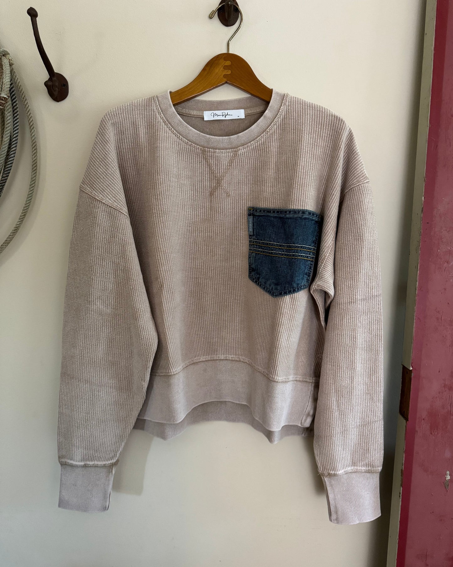 Beige sweatshirt with a blue pocket on a hanger against a white wall.