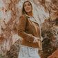 Woman in a brown jacket and white skirt standing in a rocky, natural setting.