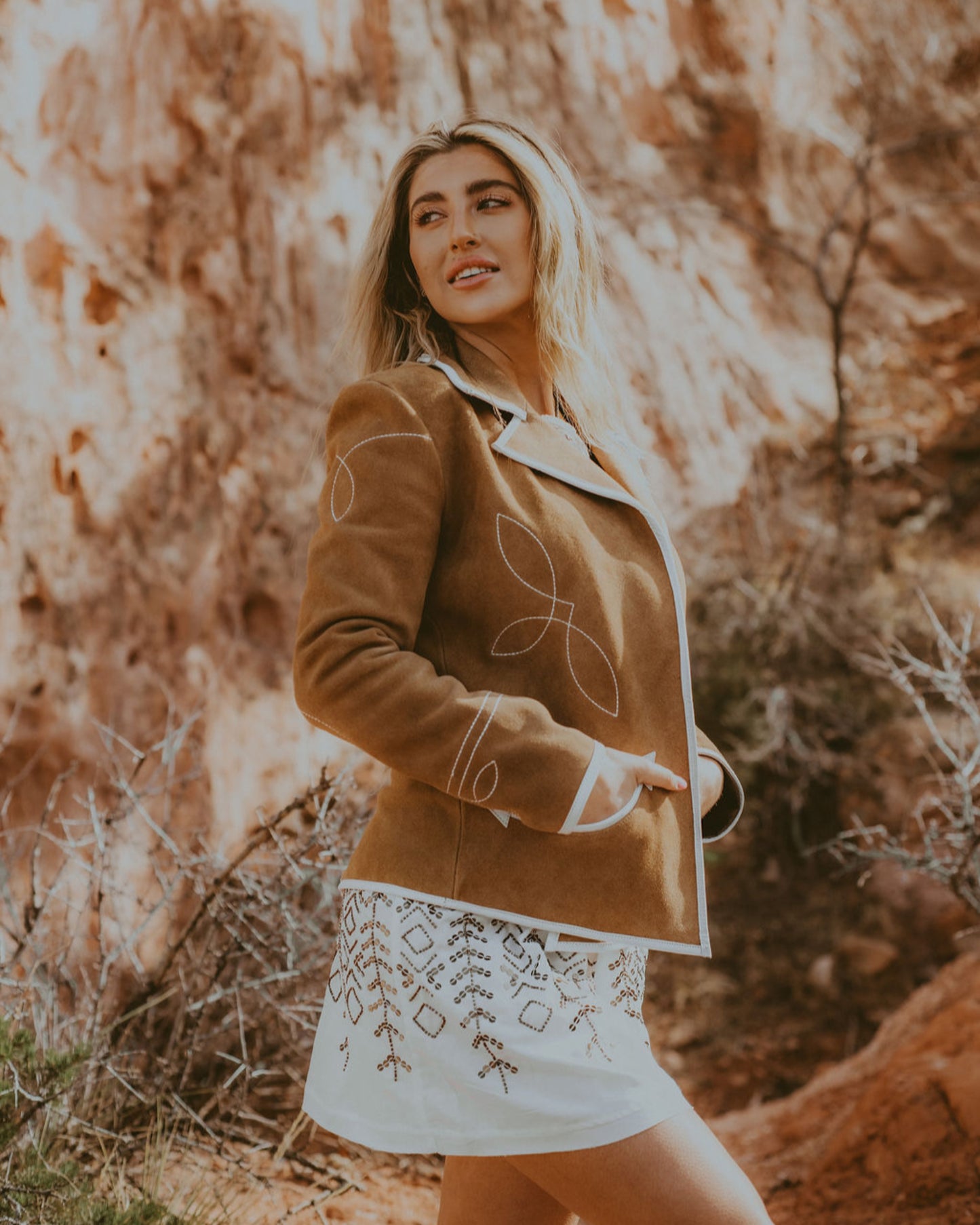 Woman in a brown jacket and white skirt standing in a rocky, natural setting.