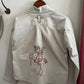 Light-colored jacket with embroidered designs on a hanger against a white wall.