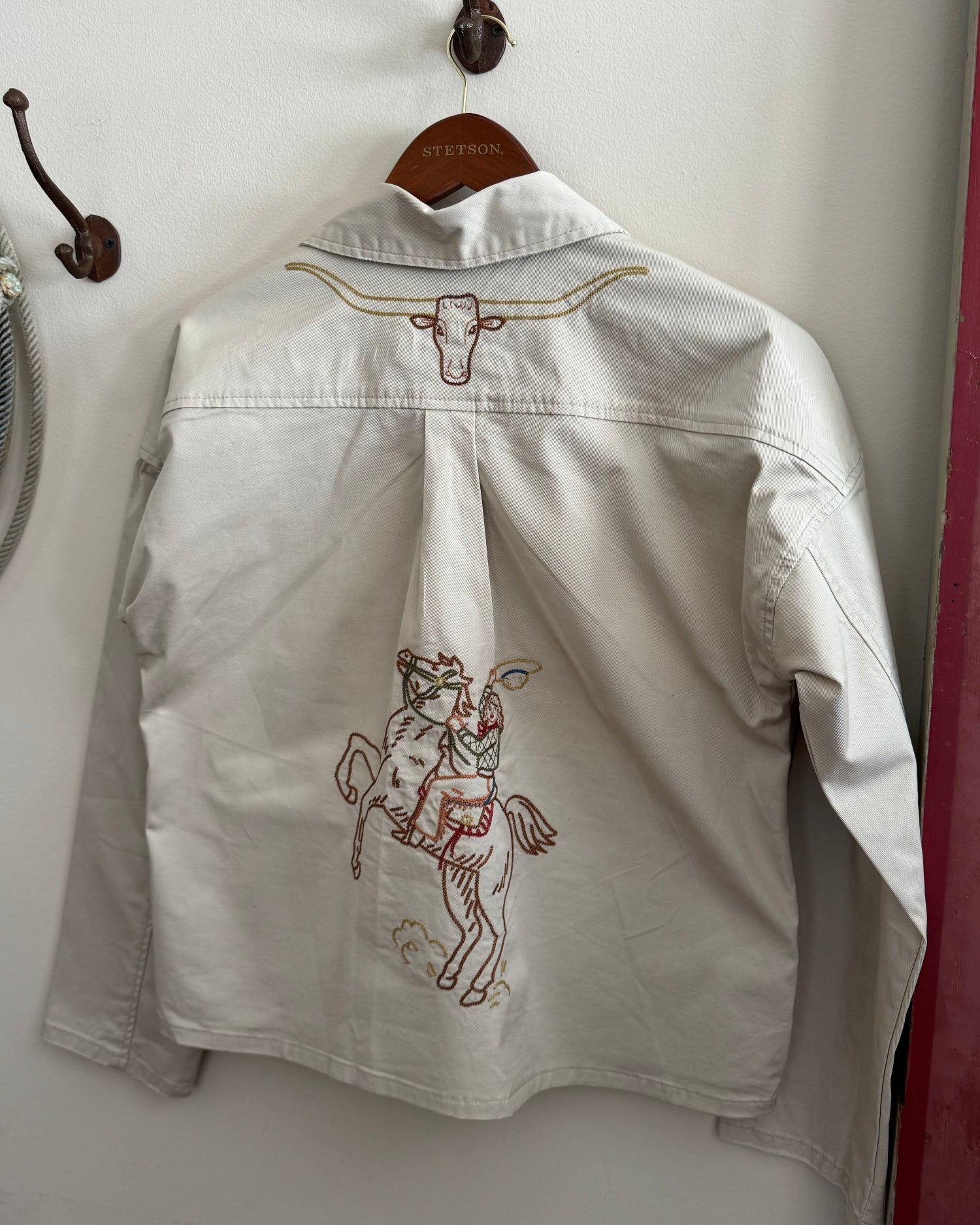 Light-colored jacket with embroidered designs on a hanger against a white wall.