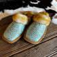 A pair of western-style slip-on slippers with turquoise tooled leather uppers, natural brown fur trim, and tan soles, displayed on a rustic wooden surface with a black-and-white cowhide rug in the background.