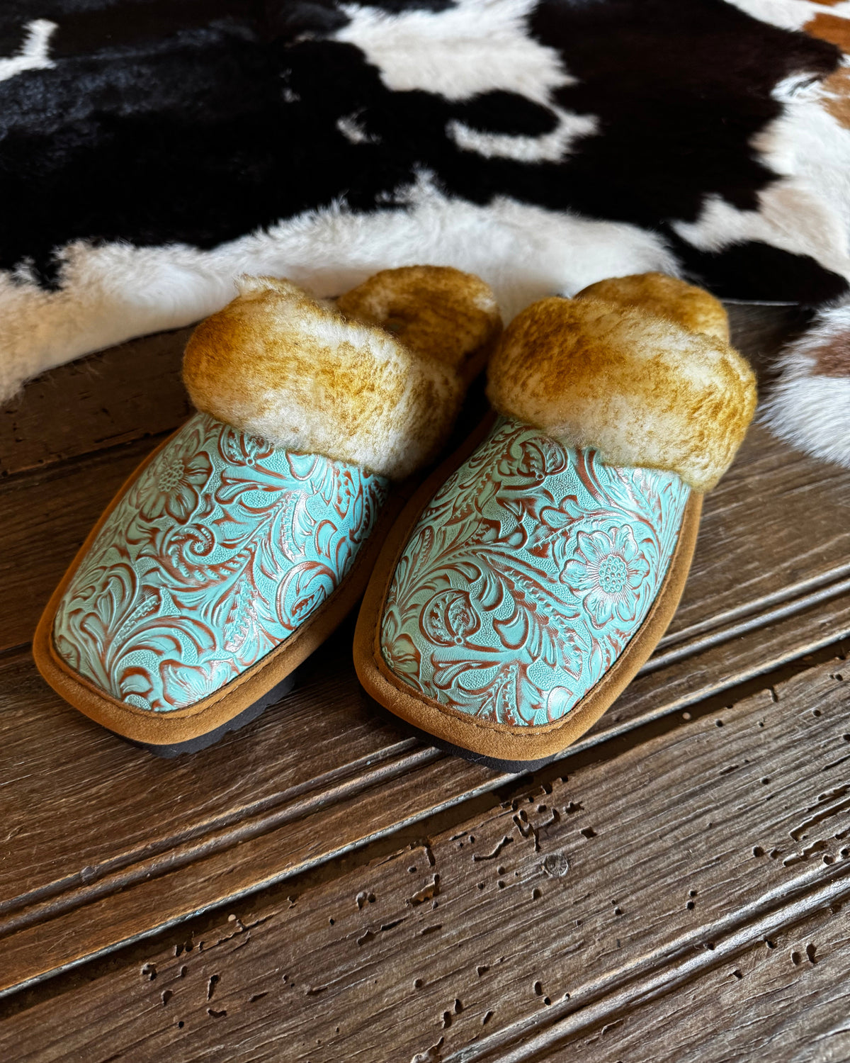 A pair of western-style slip-on slippers with turquoise tooled leather uppers, natural brown fur trim, and tan soles, displayed on a rustic wooden surface with a black-and-white cowhide rug in the background.