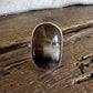 A silver ring with a large, polished oval cat's eye gemstone, displayed on a rustic wooden surface with a white faux fur trim at the top.