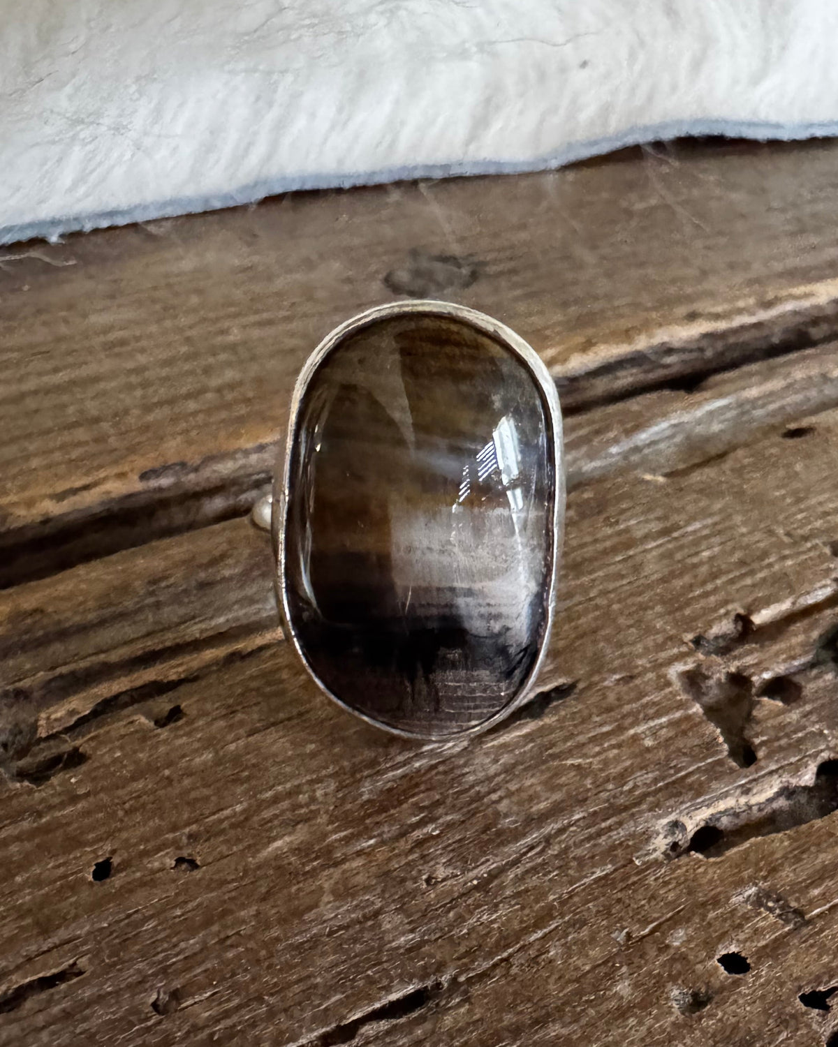A silver ring with a large, polished oval cat's eye gemstone, displayed on a rustic wooden surface with a white faux fur trim at the top.
