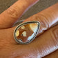 A close-up of a hand wearing a silver ring with a teardrop-shaped agate stone featuring brown and cream patterns, set against a rustic wooden background.