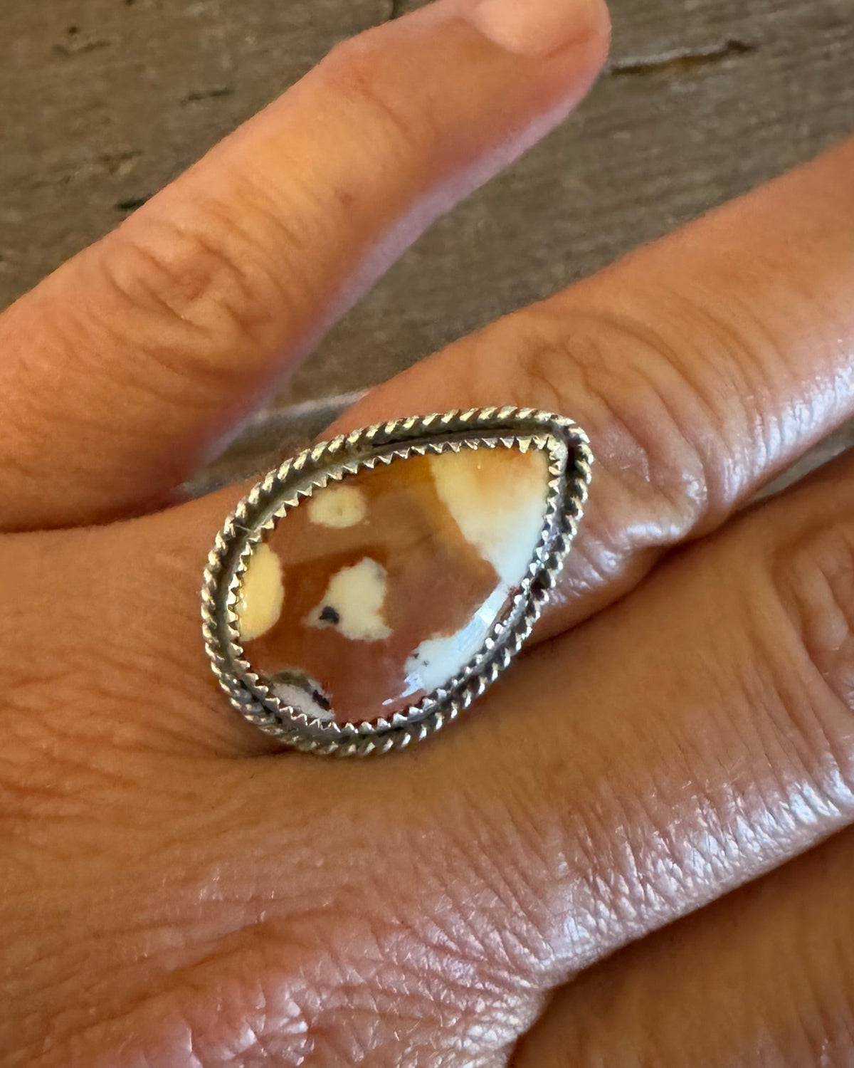 A close-up of a hand wearing a silver ring with a teardrop-shaped agate stone featuring brown and cream patterns, set against a rustic wooden background.