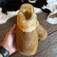 A pair of tan suede slip-on slippers with white embroidered floral patterns and a fluffy cream shearling lining, held in a hand over a rustic wooden floor with a cowhide rug.