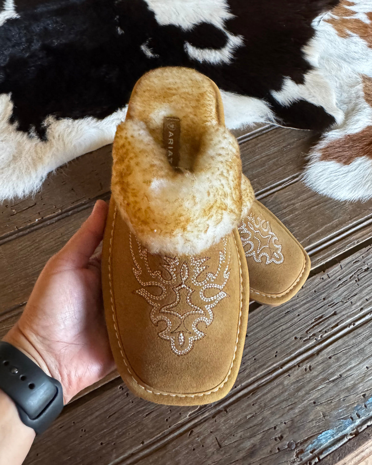 A pair of tan suede slip-on slippers with white embroidered floral patterns and a fluffy cream shearling lining, held in a hand over a rustic wooden floor with a cowhide rug.