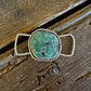 A silver-toned ring with a large, organic-shaped green stone featuring dark matrix patterns, resting on a rustic wooden surface.