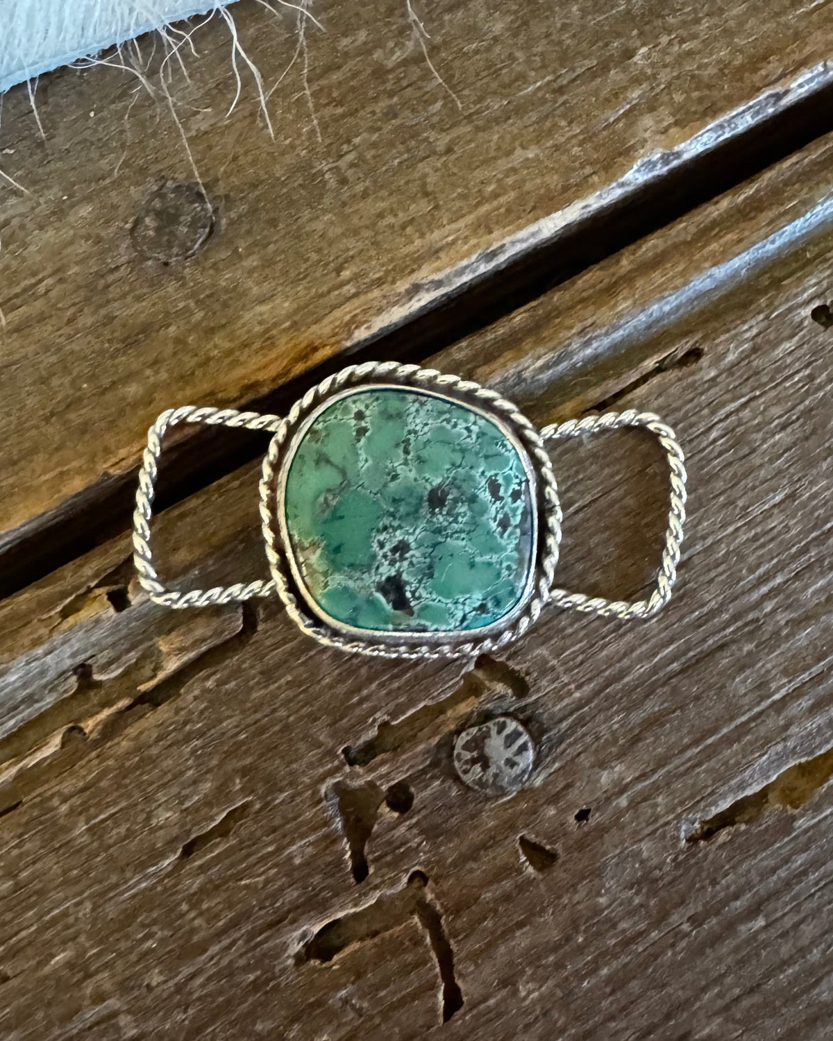 A silver-toned ring with a large, organic-shaped green stone featuring dark matrix patterns, resting on a rustic wooden surface.