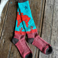 A pair of crew socks laid flat on a rustic wooden surface. The socks feature a turquoise upper section with a red cowboy and cactus pattern, a red horizontal band, and a dusty rose lower section with dark gray heels, toes, and cuffs.