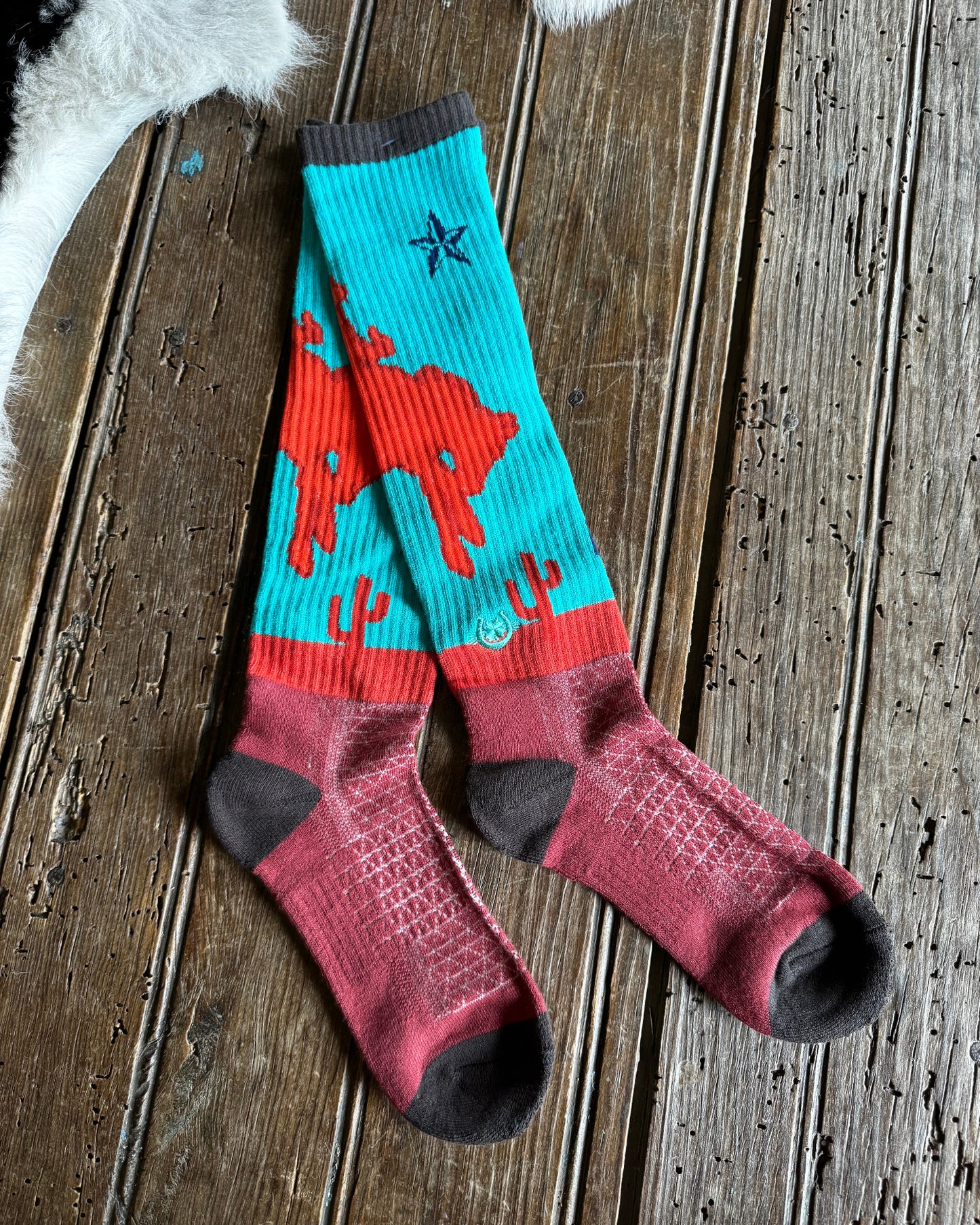 A pair of crew socks laid flat on a rustic wooden surface. The socks feature a turquoise upper section with a red cowboy and cactus pattern, a red horizontal band, and a dusty rose lower section with dark gray heels, toes, and cuffs.