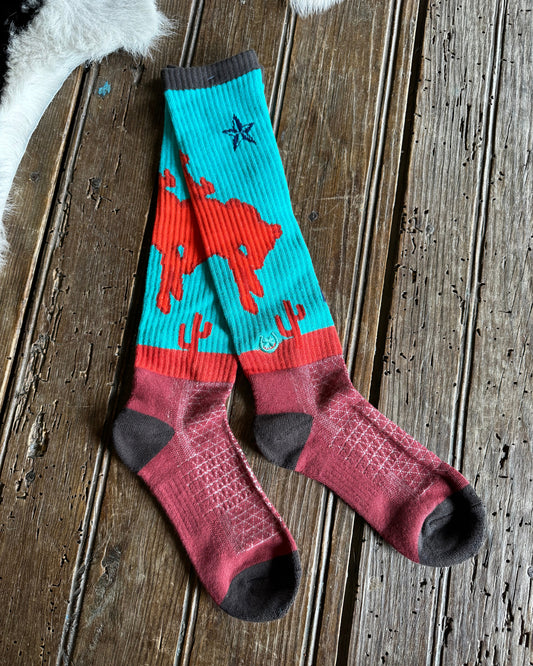 A pair of crew socks laid flat on a rustic wooden surface. The socks feature a turquoise upper section with a red cowboy and cactus pattern, a red horizontal band, and a dusty rose lower section with dark gray heels, toes, and cuffs.