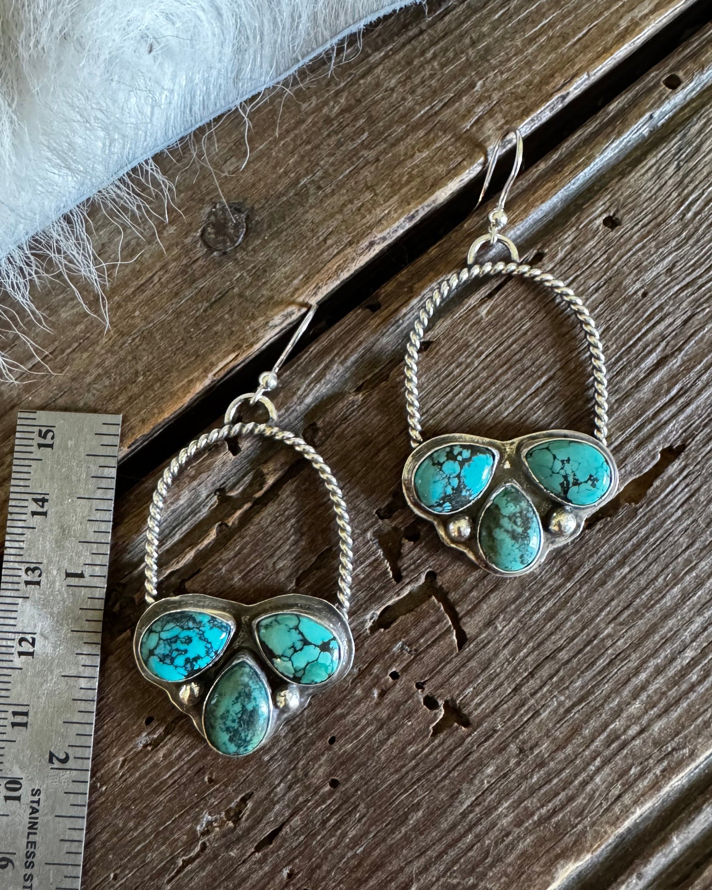 A pair of silver hoop earrings with heart-shaped clusters of turquoise stones, displayed on a rustic wooden surface next to a metal ruler for scale.