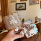 A hand holds two western-themed leather hair clips with white plastic spring clips, one featuring a white cowboy illustration and the other a western landscape print, against a rustic indoor background.