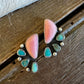 A pair of handmade turquoise and pink stone stud earrings with silver settings, displayed on a rustic dark wood surface next to a white faux fur trim.