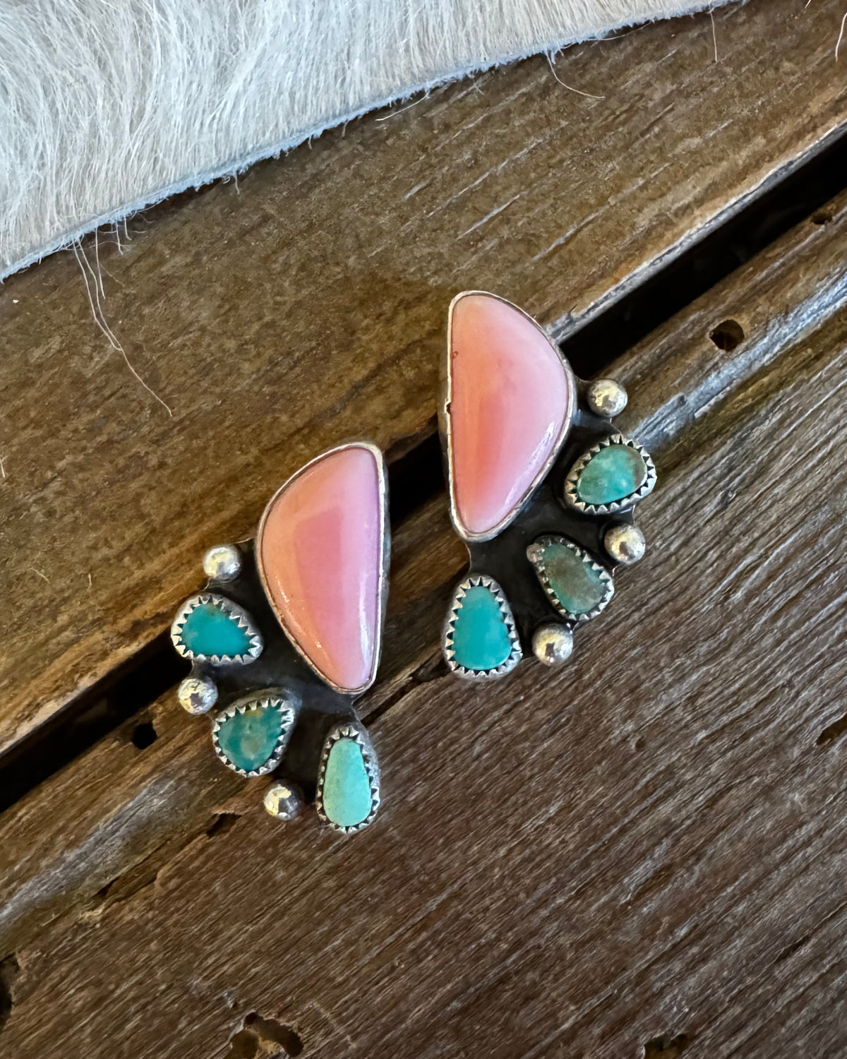 A pair of handmade turquoise and pink stone stud earrings with silver settings, displayed on a rustic dark wood surface next to a white faux fur trim.