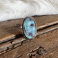 A silver-toned ring with an oval turquoise stone resting on a rustic wooden surface, with white faux fur and a dark blanket in the background.