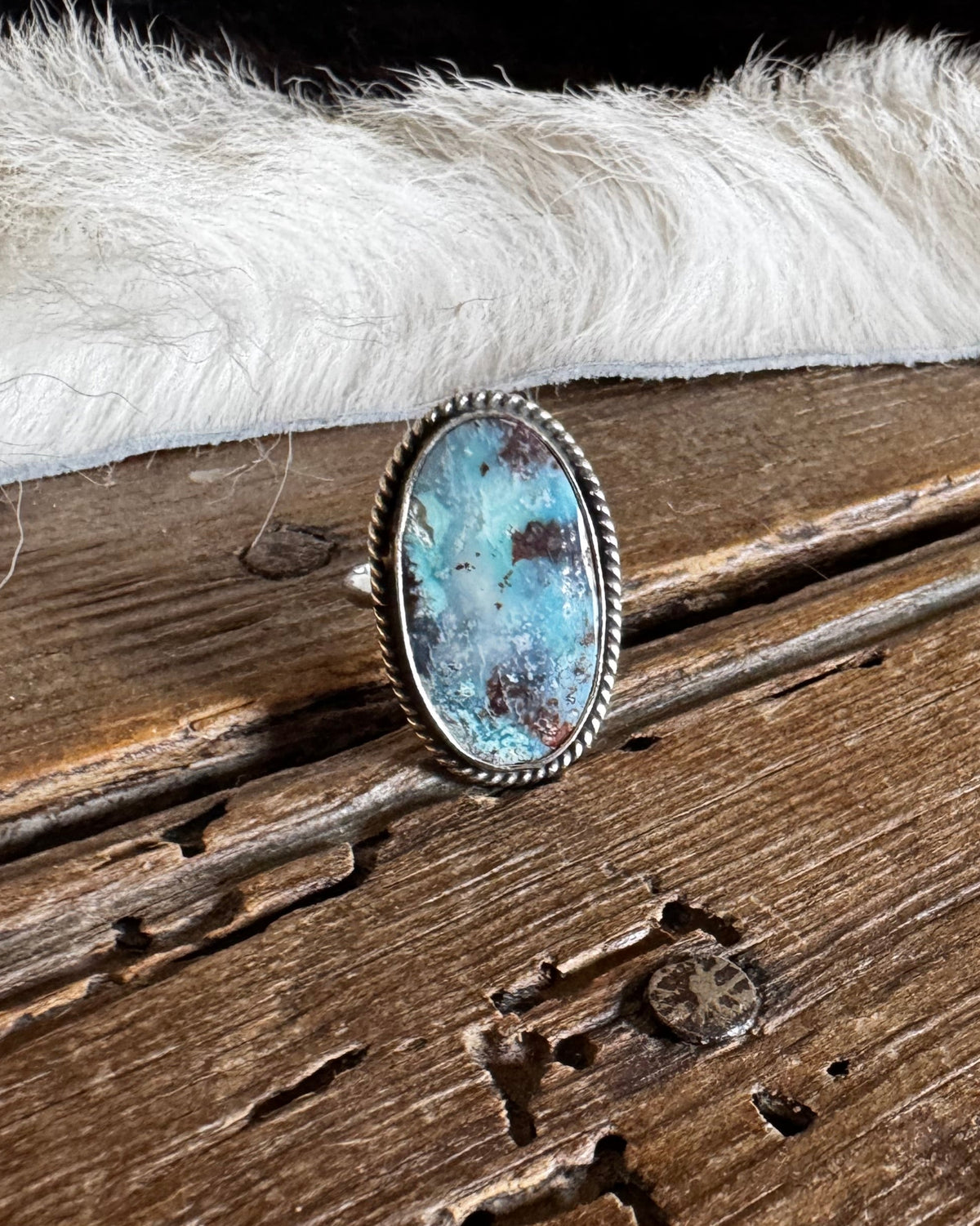 A silver-toned ring with an oval turquoise stone resting on a rustic wooden surface, with white faux fur and a dark blanket in the background.