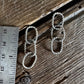 A pair of small silver-toned woven rope earrings laid on a rustic wooden surface next to a stainless steel ruler for scale.