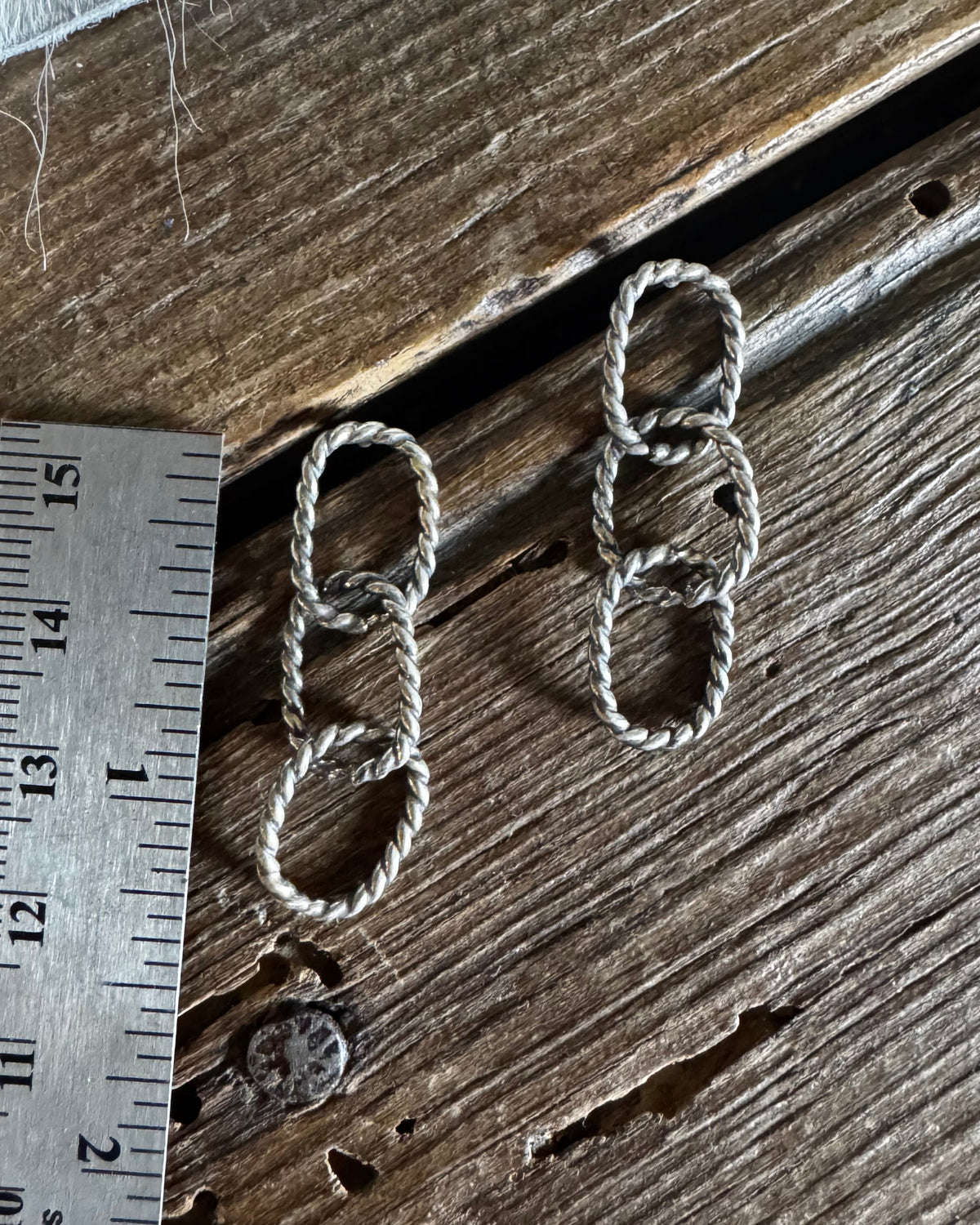 A pair of small silver-toned woven rope earrings laid on a rustic wooden surface next to a stainless steel ruler for scale.