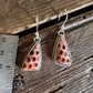 A pair of handmade earrings with coral-colored stone pendants in silver settings, displayed on a rustic wooden surface next to a metal ruler for scale.
