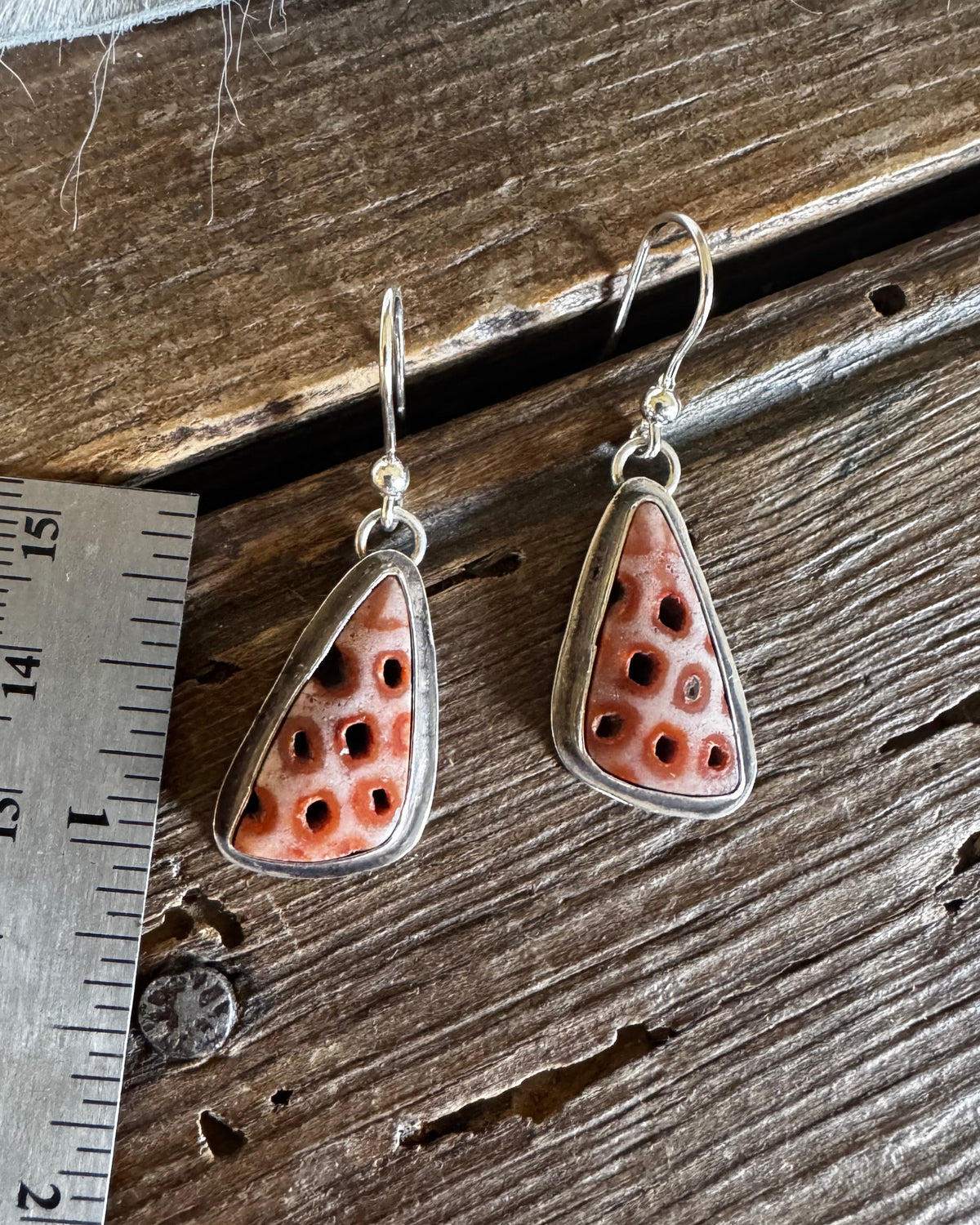 A pair of handmade earrings with coral-colored stone pendants in silver settings, displayed on a rustic wooden surface next to a metal ruler for scale.