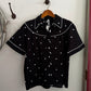 Black shirt with white polka dots on a hanger against a white wall.