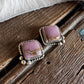 A pair of silver-toned statement earrings with square pinkish-purple stone inlays, resting on a rustic dark wood surface with a white faux-fur texture in the background.