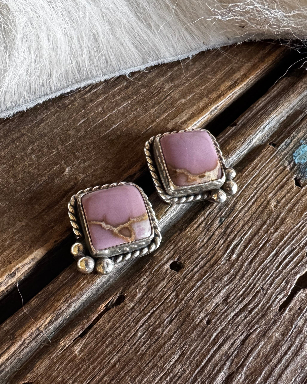 A pair of silver-toned statement earrings with square pinkish-purple stone inlays, resting on a rustic dark wood surface with a white faux-fur texture in the background.