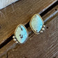 A pair of gold-tone stud earrings with irregular light blue-green turquoise stones and small bead drops, displayed on a rustic wooden surface with a white faux-fur trim at the top.