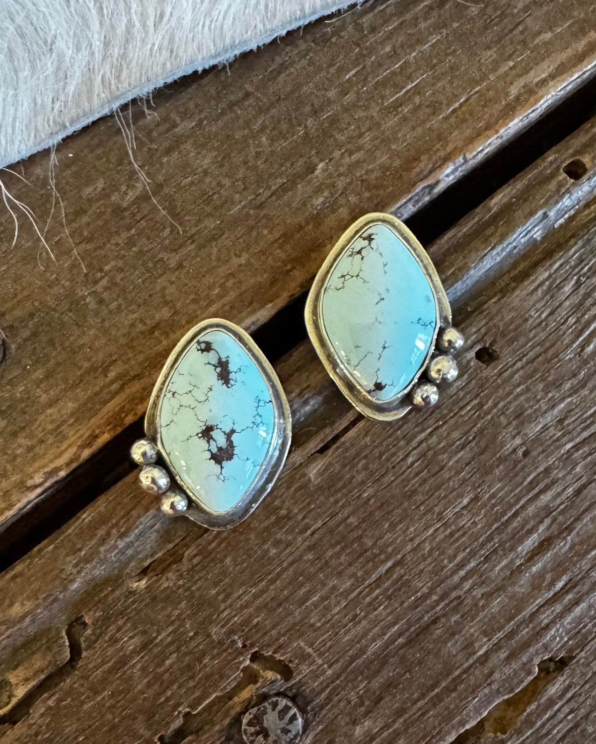 A pair of gold-tone stud earrings with irregular light blue-green turquoise stones and small bead drops, displayed on a rustic wooden surface with a white faux-fur trim at the top.