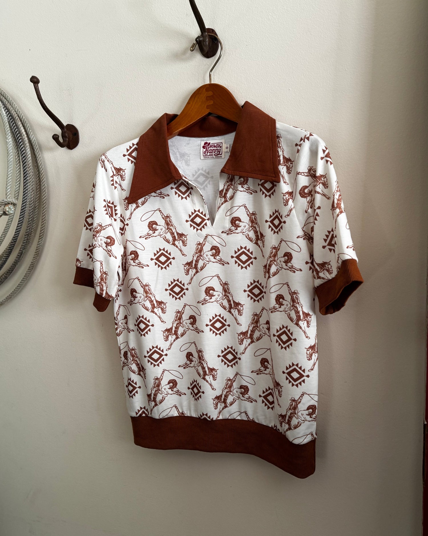 White shirt with brown collar and cuffs hanging on a wooden hanger against a white wall.