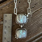 A silver link necklace with two square iridescent stones set in a twisted silver frame, displayed next to a metal ruler on a rustic wooden surface.