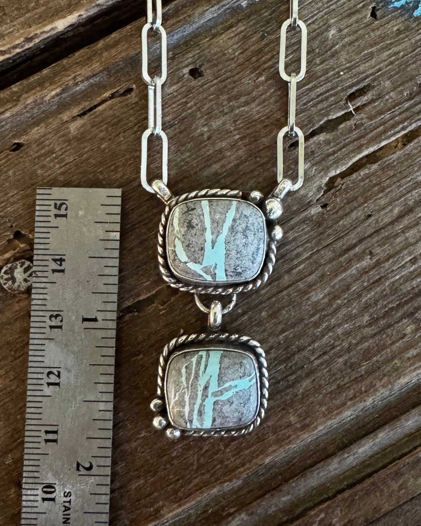 A silver link necklace with two square iridescent stones set in a twisted silver frame, displayed next to a metal ruler on a rustic wooden surface.