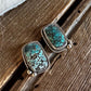 A pair of rectangular turquoise stud earrings with silver-toned bezel settings, resting on a rustic wooden surface with a white fur trim visible at the top.