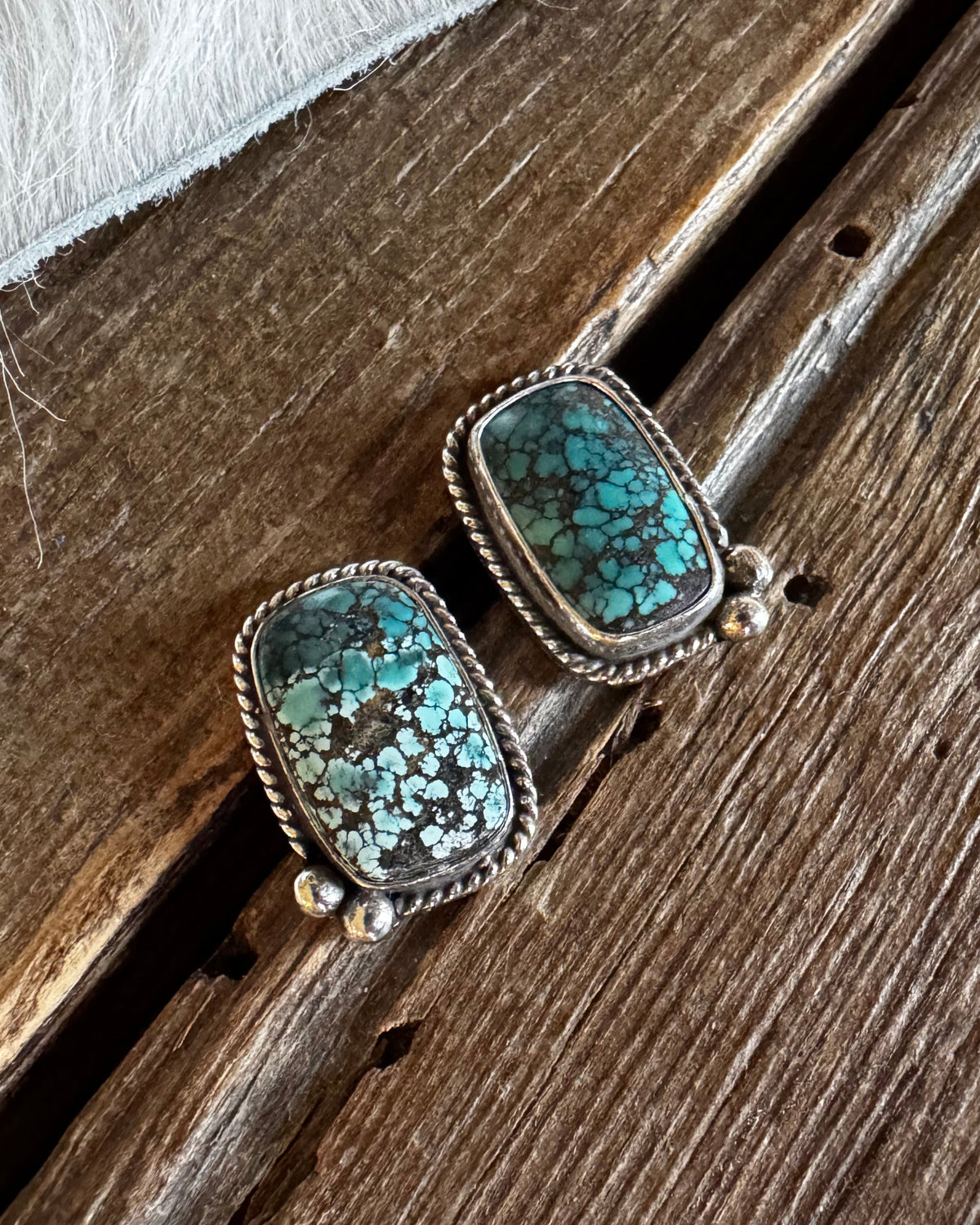 A pair of rectangular turquoise stud earrings with silver-toned bezel settings, resting on a rustic wooden surface with a white fur trim visible at the top.