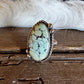 A silver ring featuring a large oval turquoise stone with black matrix veining, displayed on a rustic wooden surface with a white fur trim in the background.