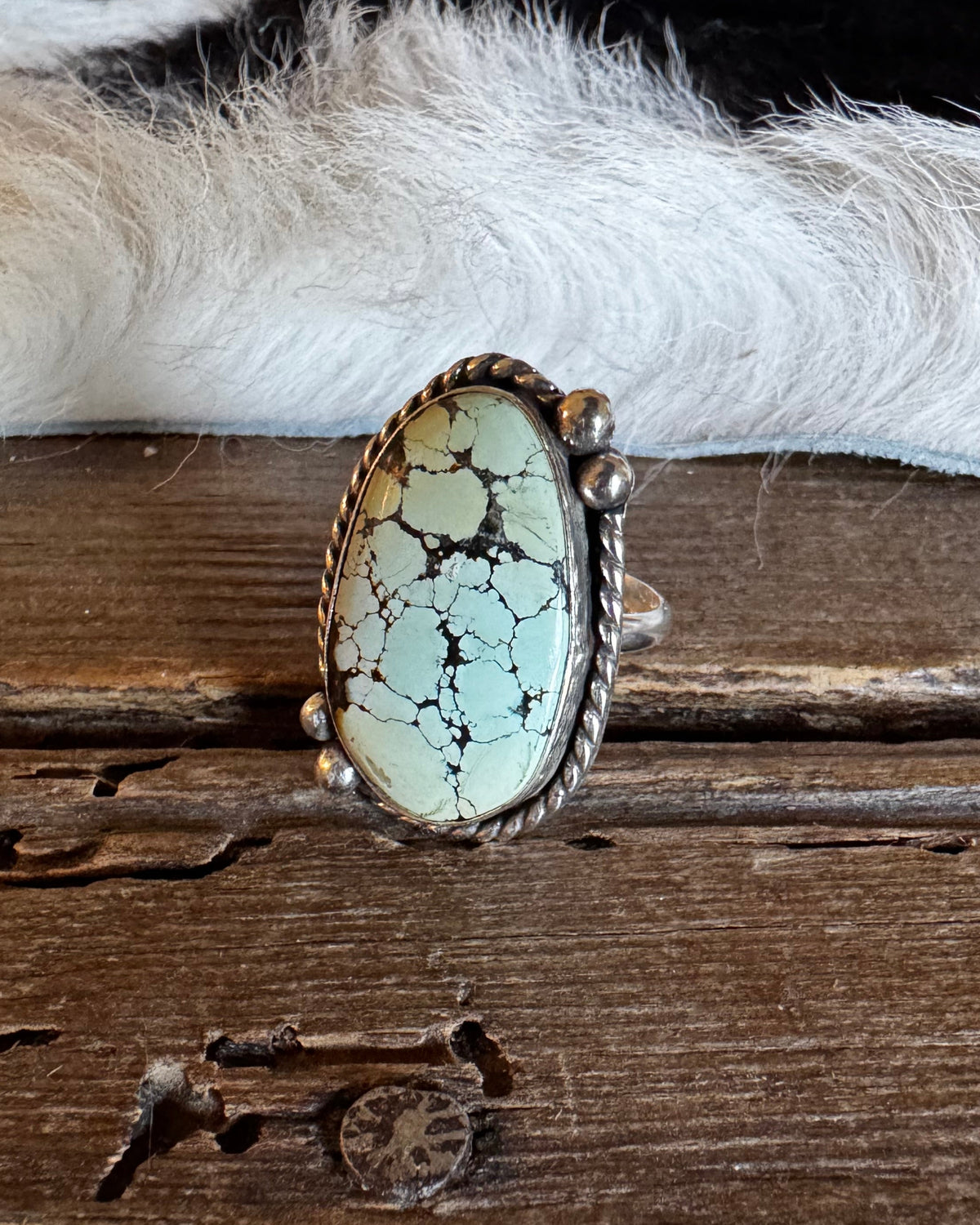 A silver ring featuring a large oval turquoise stone with black matrix veining, displayed on a rustic wooden surface with a white fur trim in the background.