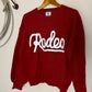 Red sweater with 'Rodeo' text on a wooden hanger against a white wall.