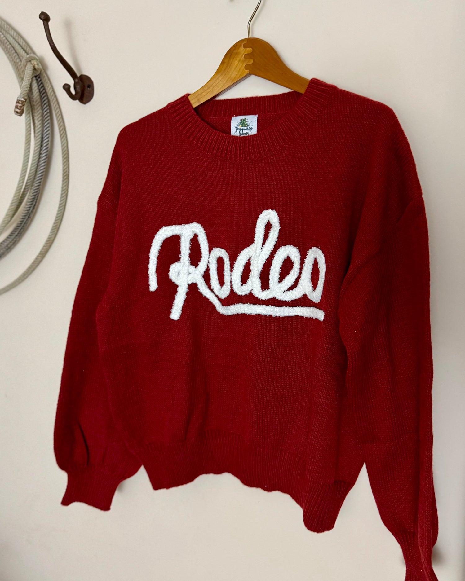 Red sweater with 'Rodeo' text on a wooden hanger against a white wall.