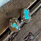 A pair of silver stud earrings with teardrop-shaped turquoise and brown stone inlays, resting on a rustic wooden surface next to white faux fur.