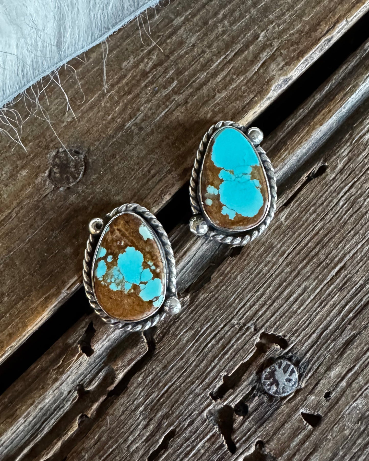 A pair of silver stud earrings with teardrop-shaped turquoise and brown stone inlays, resting on a rustic wooden surface next to white faux fur.