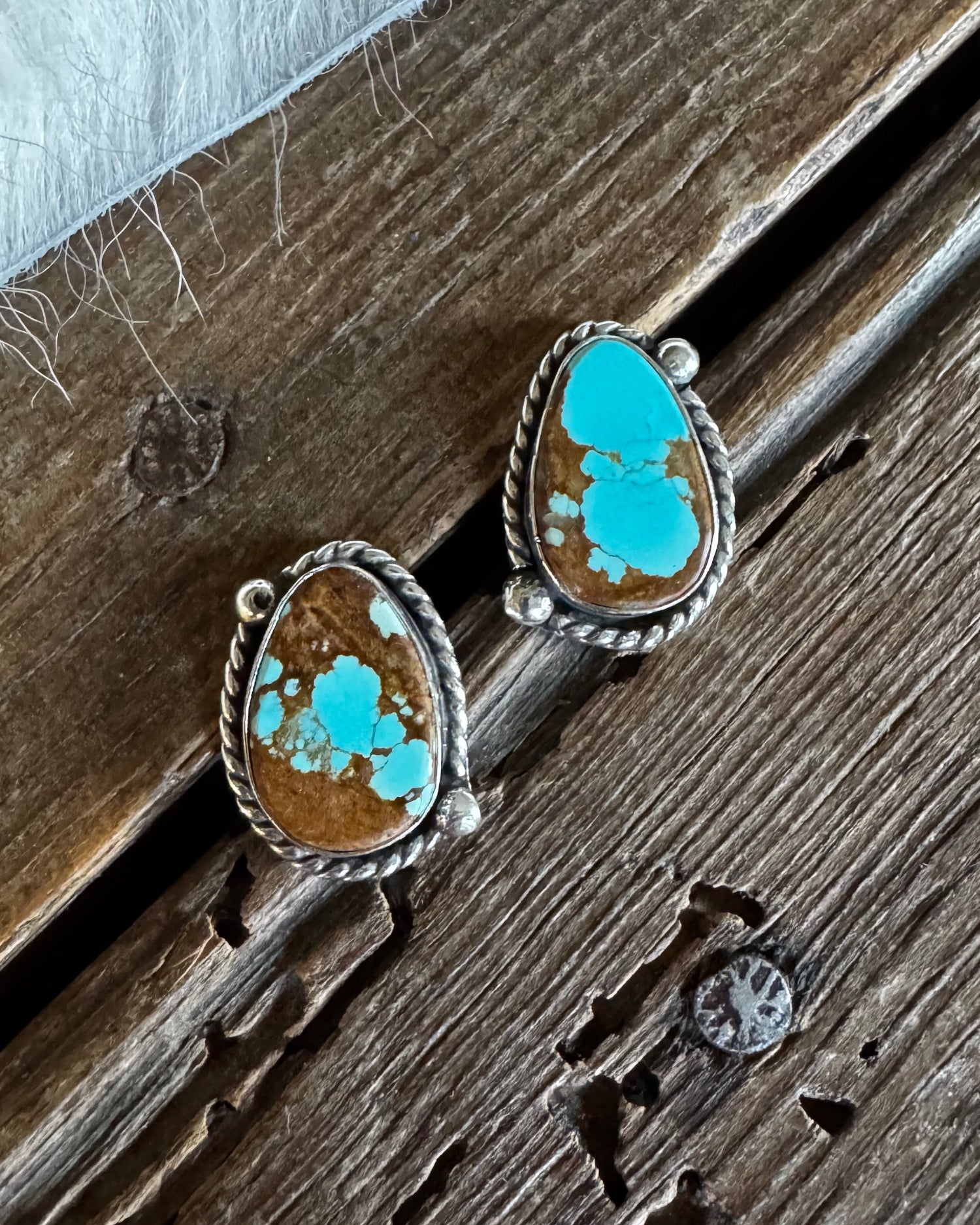 A pair of silver stud earrings with teardrop-shaped turquoise and brown stone inlays, resting on a rustic wooden surface next to white faux fur.