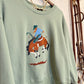 A sage green crewneck sweatshirt hanging on a wooden hanger, featuring a colorful embroidered cowboy riding a horse graphic on the front. The sweatshirt is styled with a gold chain necklace, set against a rustic wall with leather straps hanging on the side.