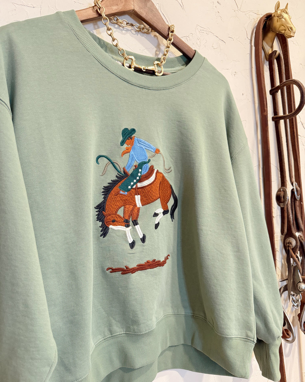 A sage green crewneck sweatshirt hanging on a wooden hanger, featuring a colorful embroidered cowboy riding a horse graphic on the front. The sweatshirt is styled with a gold chain necklace, set against a rustic wall with leather straps hanging on the side.