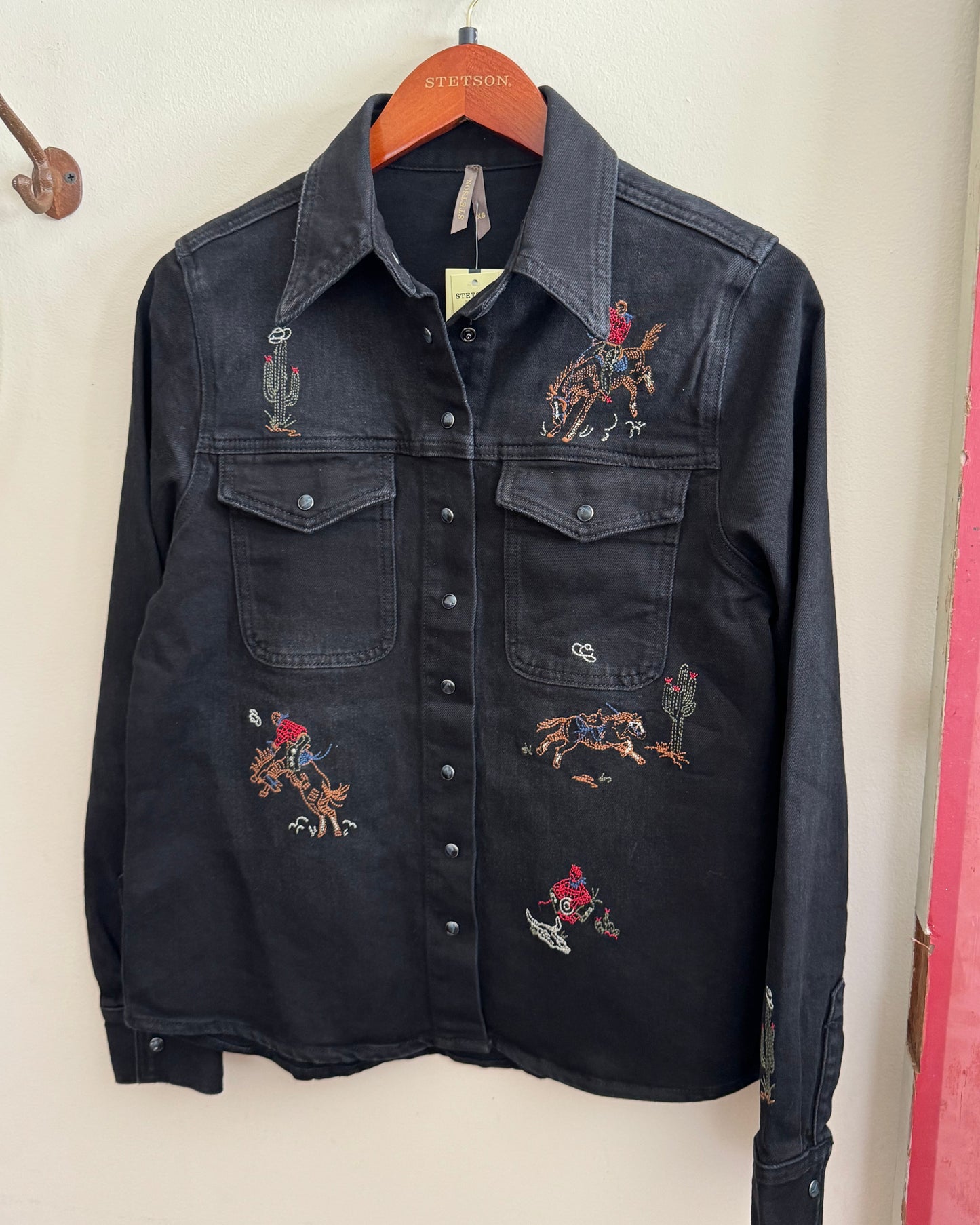 Black denim shirt with embroidered designs on a hanger against a white wall.