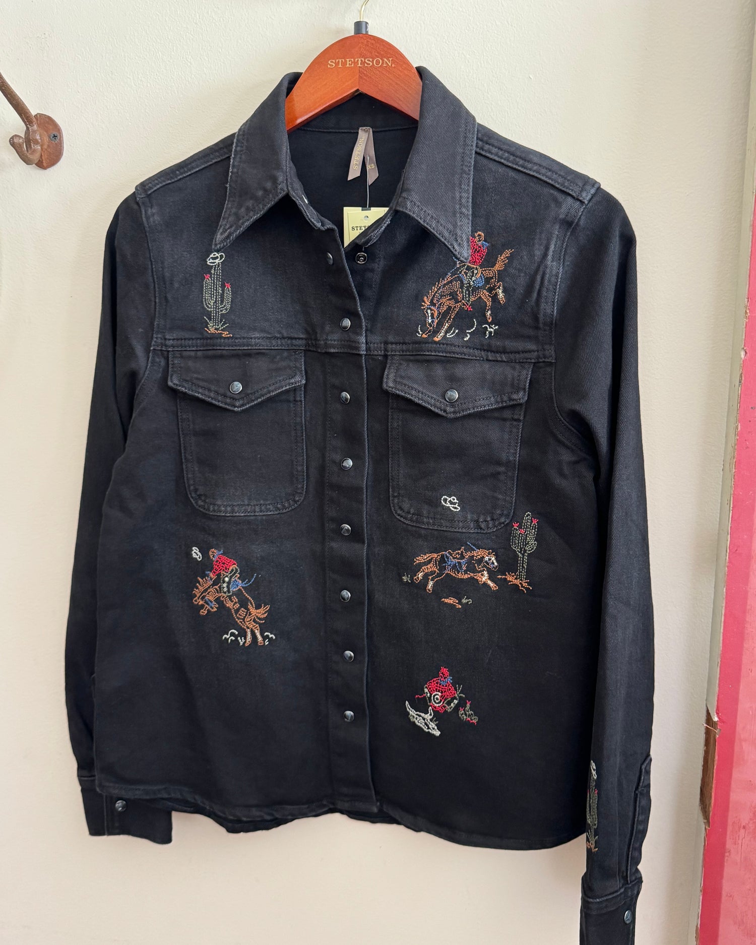 Black denim shirt with embroidered designs on a hanger against a white wall.
