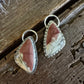A pair of triangular drop earrings with pinkish-brown and cream marbled cabochon stones in silver bezel settings, displayed on a rustic dark wood surface.